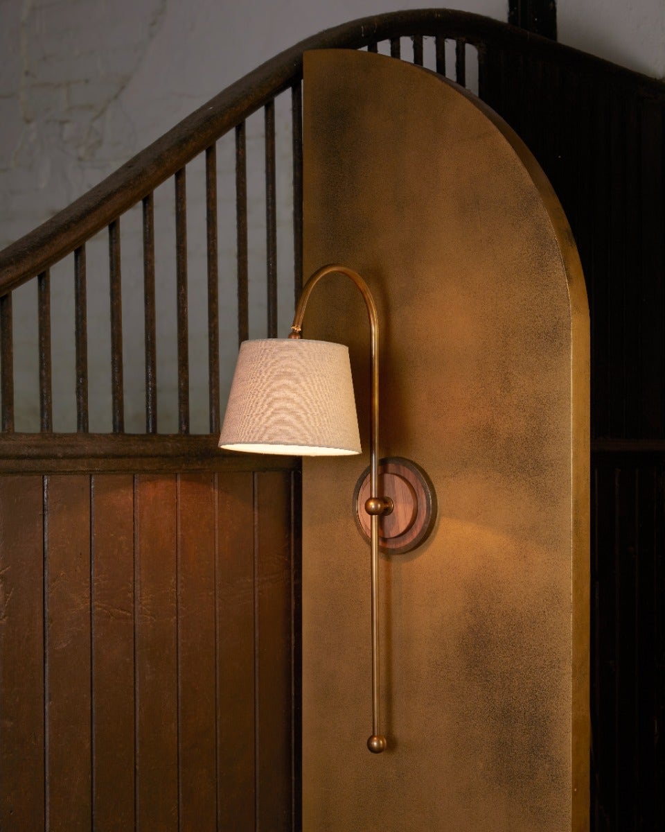 Arched brass and walnut wall light with linen shade on brass arch board in stable setting.